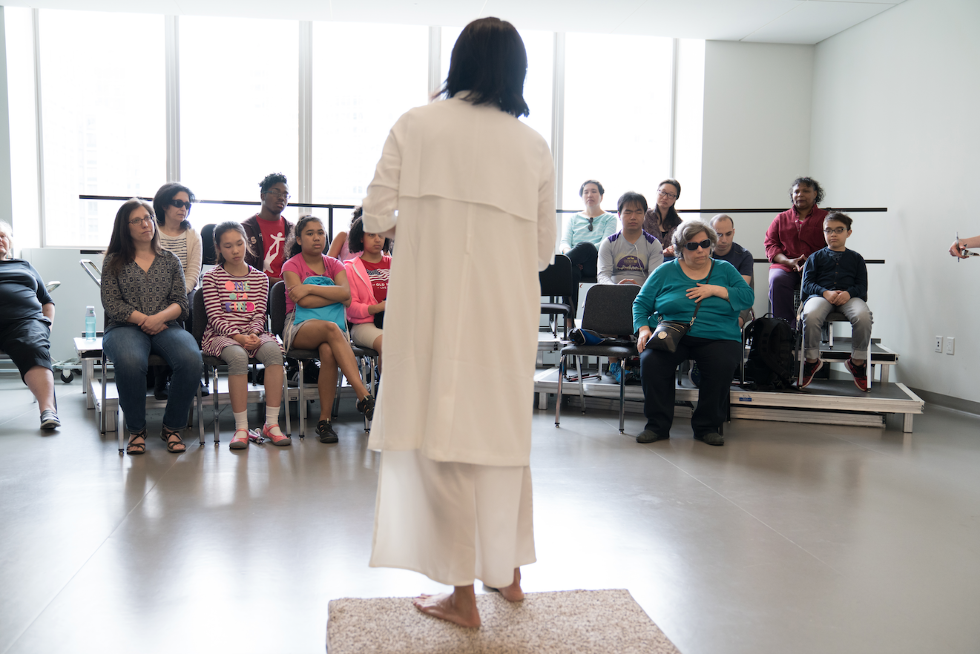 Dance artist Mana Hashimoto is giving a lecture to a group of visually-impaired audience members. She is standing on a platform, wearing a long white dress as the audience listens.