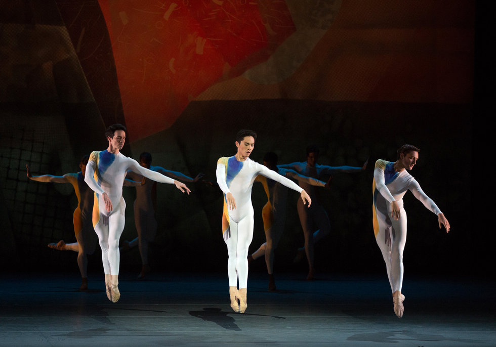Three men in colored unitards jump in unison, legs together in fifth position, arms reaching diagonally down towards their sides