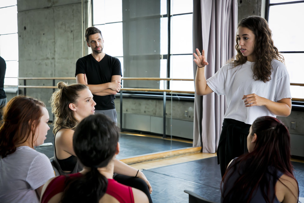 One dancer speaks to a group sitting in chairs, while a teacher looks on