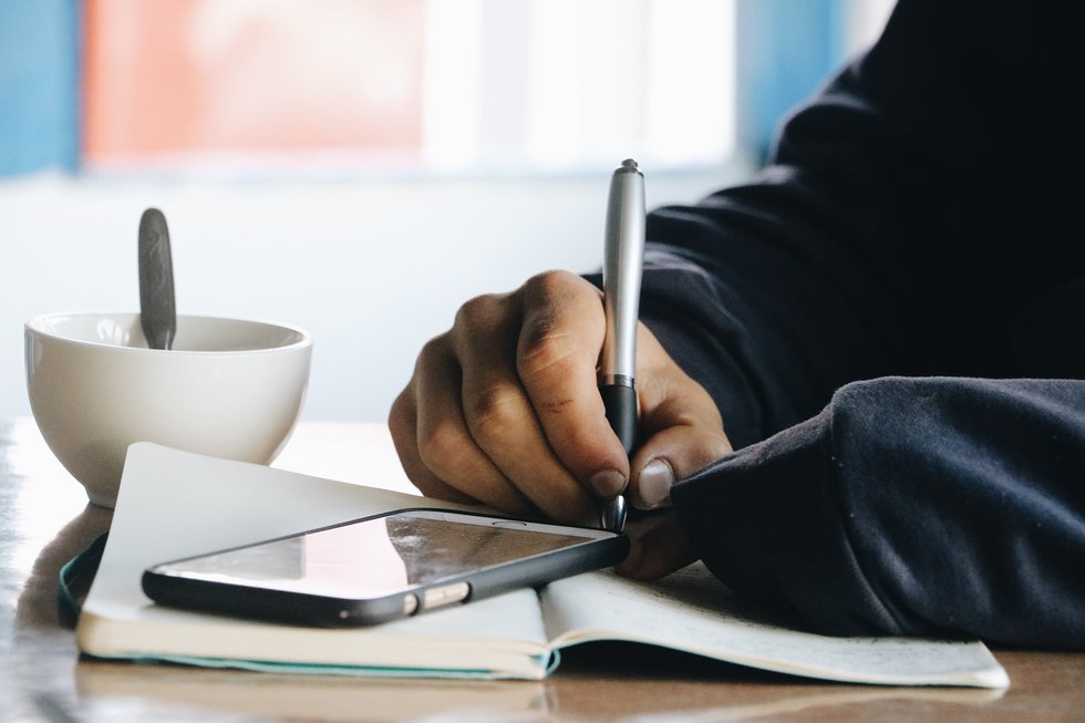 Hands writing in a notebook, with a cell phone and coffee on the table