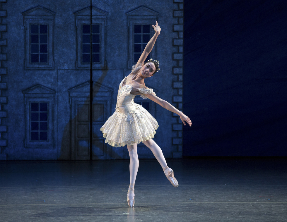 A female dancer stands onstage, balancing on one leg with her other leg raised behind her. She is wearing a white tutu and crown.