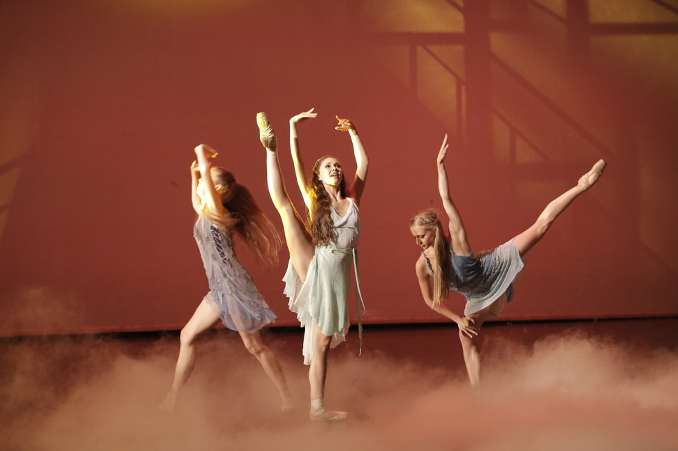 A young ballerina stands center stage, lifting her right leg high to the side and reaching both arms up in fifth position. A female dancer to her right is running past her and another dancer to her left is in arabesque.