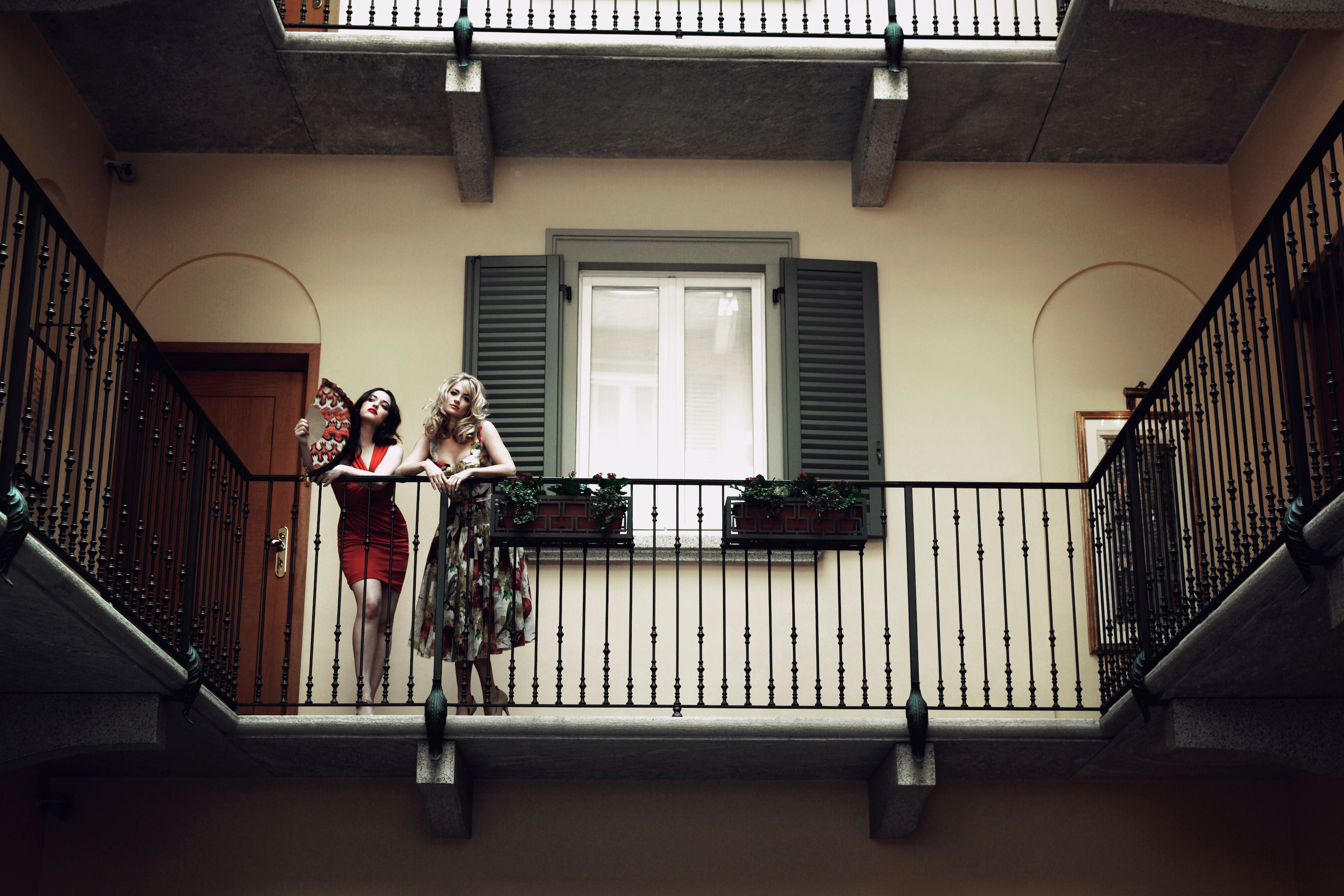 Kat Dennings og Beth Behrs i cocktailkjoler på en balkon i Milano