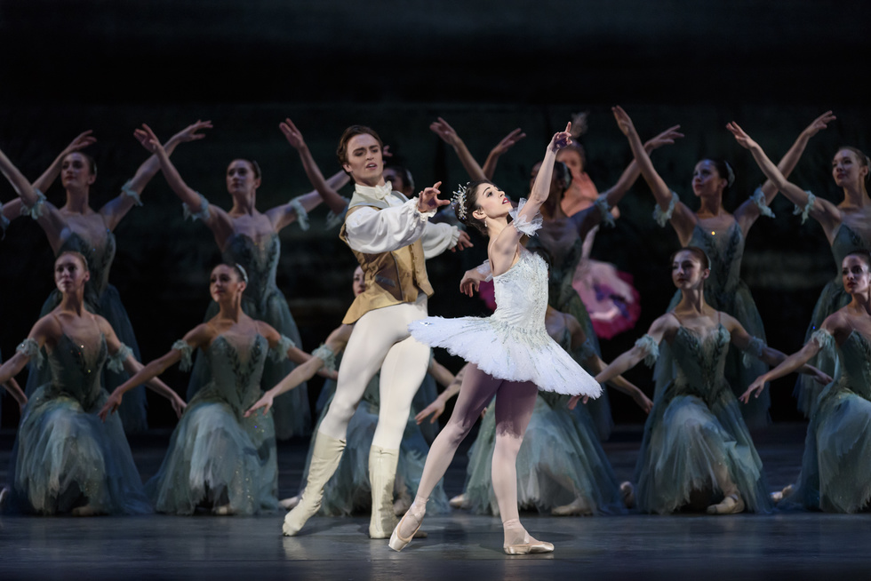 Yasmine Naghdi wearing a pale blue tutu stands in front of a group of women dressed in sheer blue dresses. She reaches towards the sky, standing in a back tendu. Behind her, Matthew Ball, dressed in white tights, a white shirt and a gold vest reaches towards her.