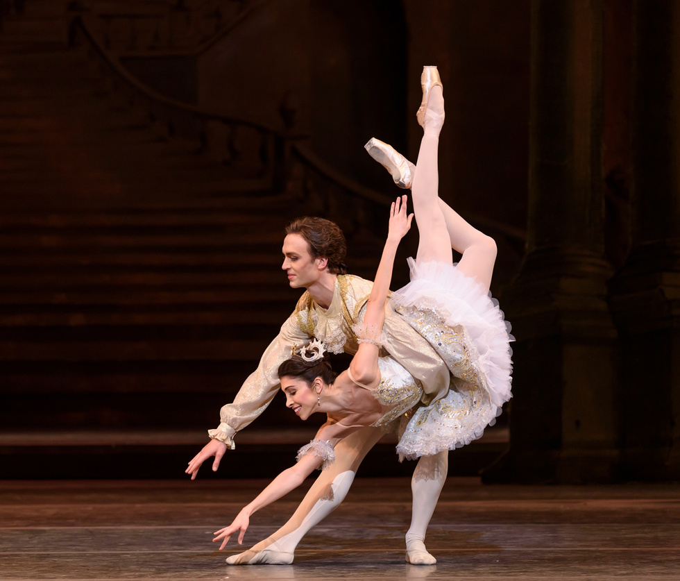 Yasmine and Naghdi are in a classic fish dive. They're both dressed in the Sleeping Beauty wedding attire. Naghdi wears a gold and white intricate tutu and Ball wears a gold vest, lace cravate and white tights.