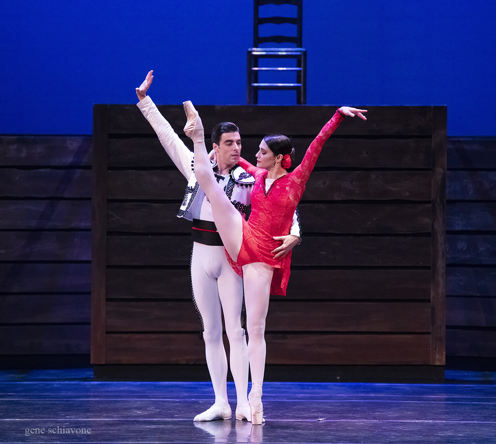Sarah Lane, wearing a short red, lacy costume, kicks her right leg high in the air. Luis Ribagorda, wearing white tights and a white bolero jacket, holds on to her waist.