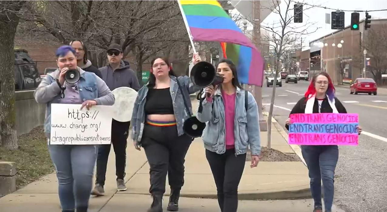 A group of people protesting while holding gay pride flags and a sign in the trans pride colors that reads "End trans genocide"