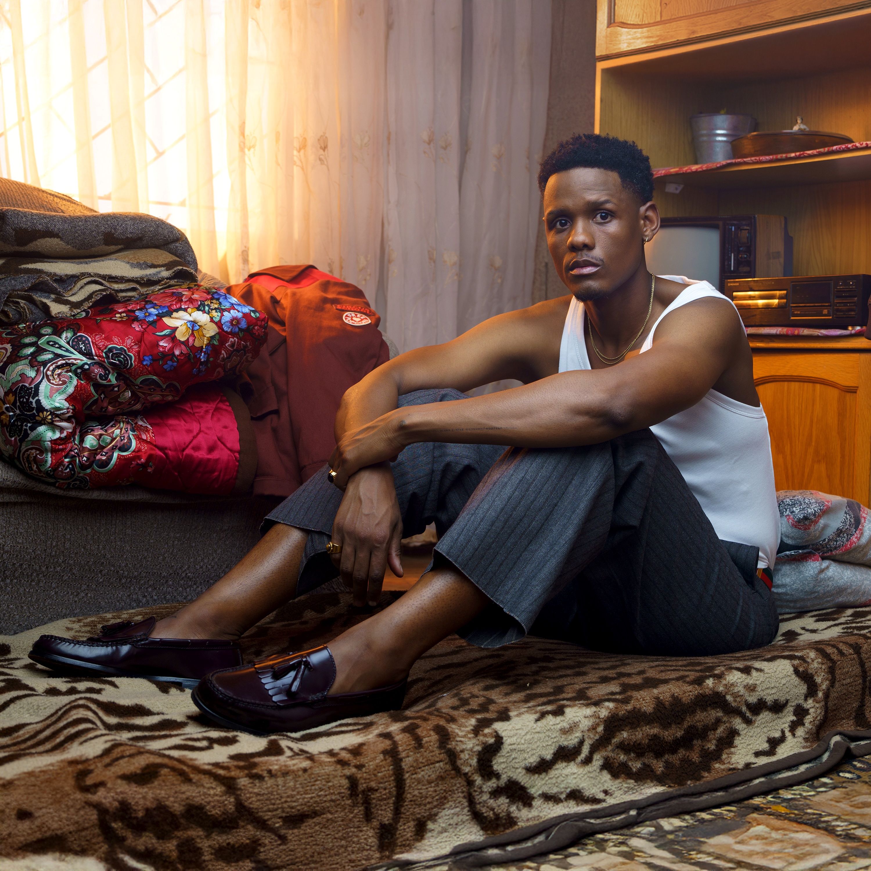 Samthing Soweto poses for the camera while seated on a blanket. He wears maroon  Dakota shoes, grey pants and a white vest.