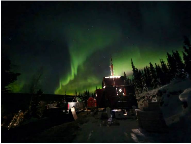 A dimly lit cabin and trees under vibrant green northern lights in a snowy, nighttime landscape.