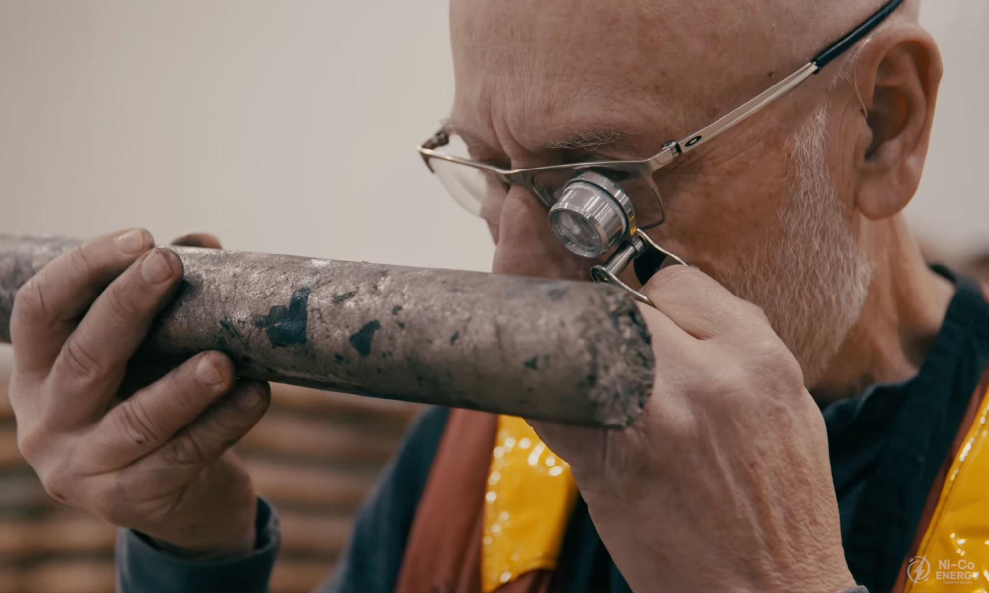 A person closely examines Ni-Co Energy cylindrical rock sample with a magnifying eyepiece while wearing glasses and a yellow vest.
