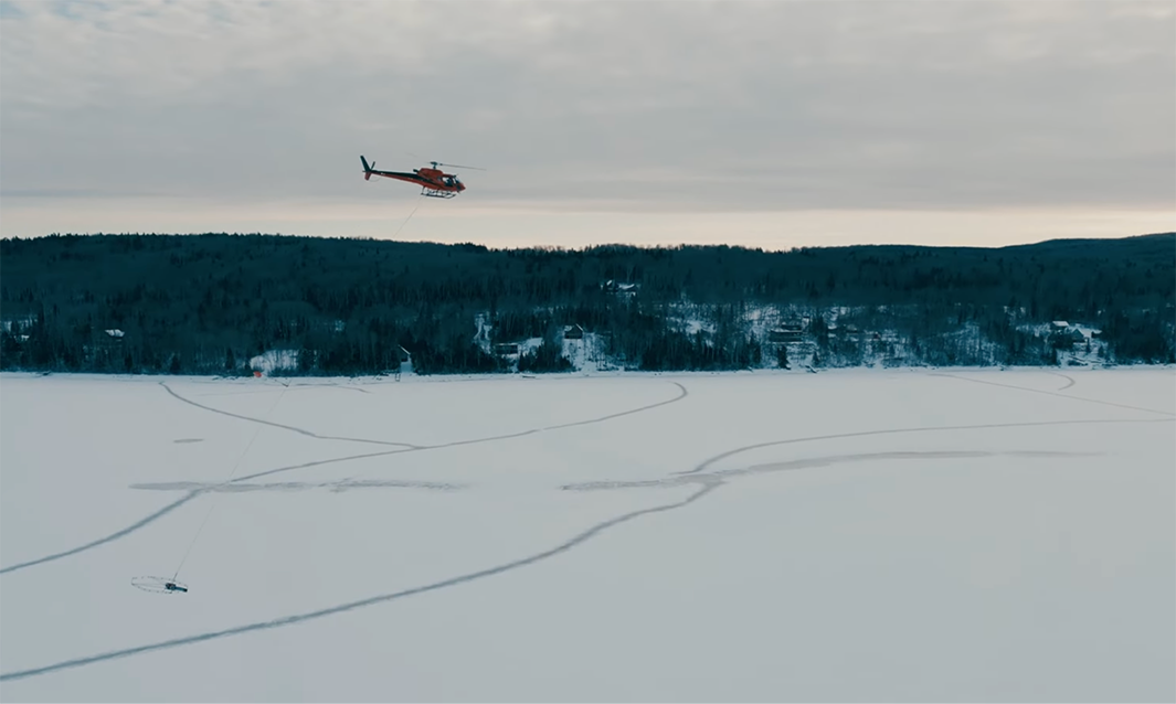 Ni-Co Energy's helicopter hovers above a snowy field with dark forested hills in the background under a cloudy sky.